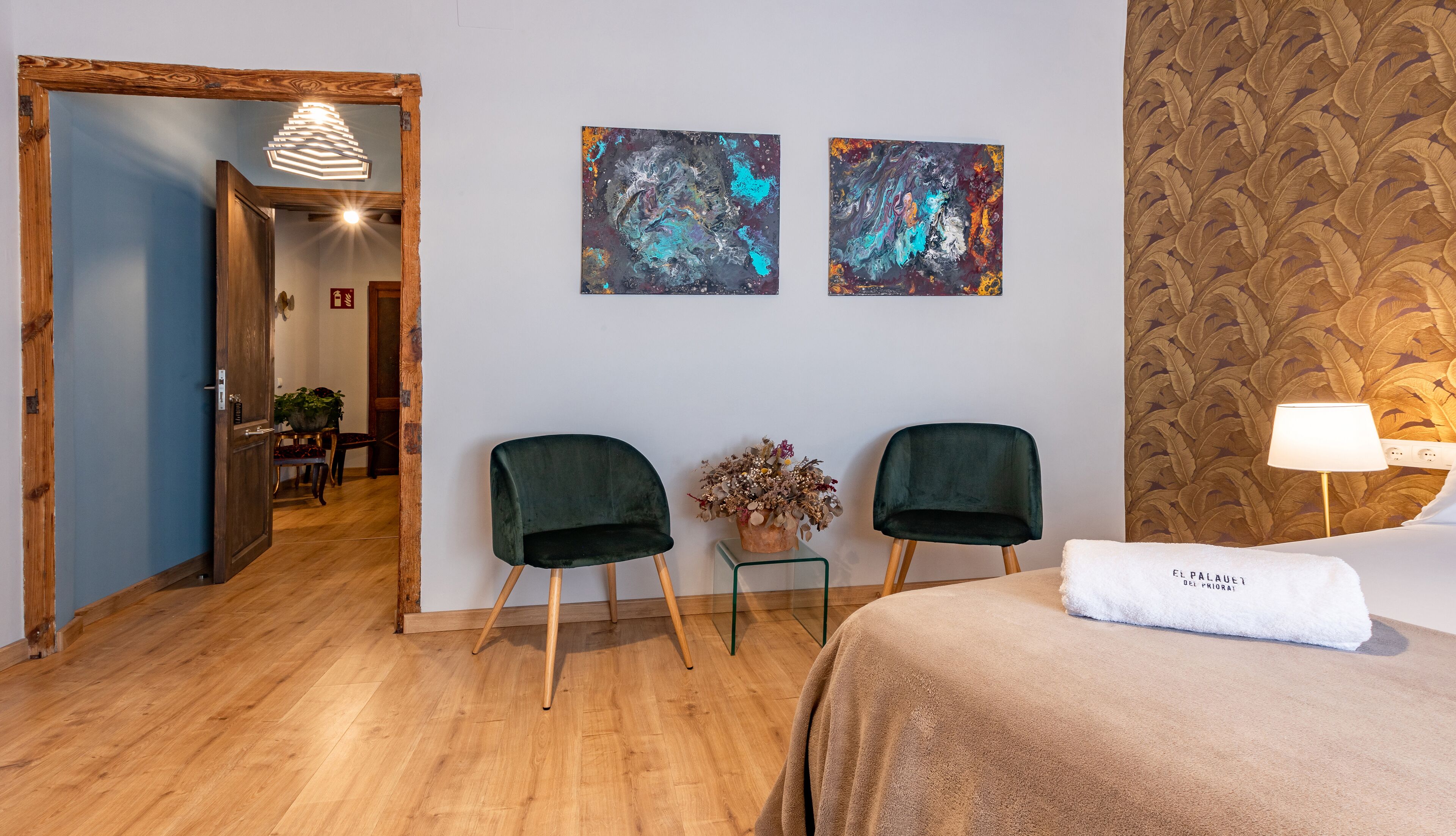 Habitación de hotel con piso de madera, dos sillas verdes, una cama con toalla blanca y dos cuadros abstractos en la pared.