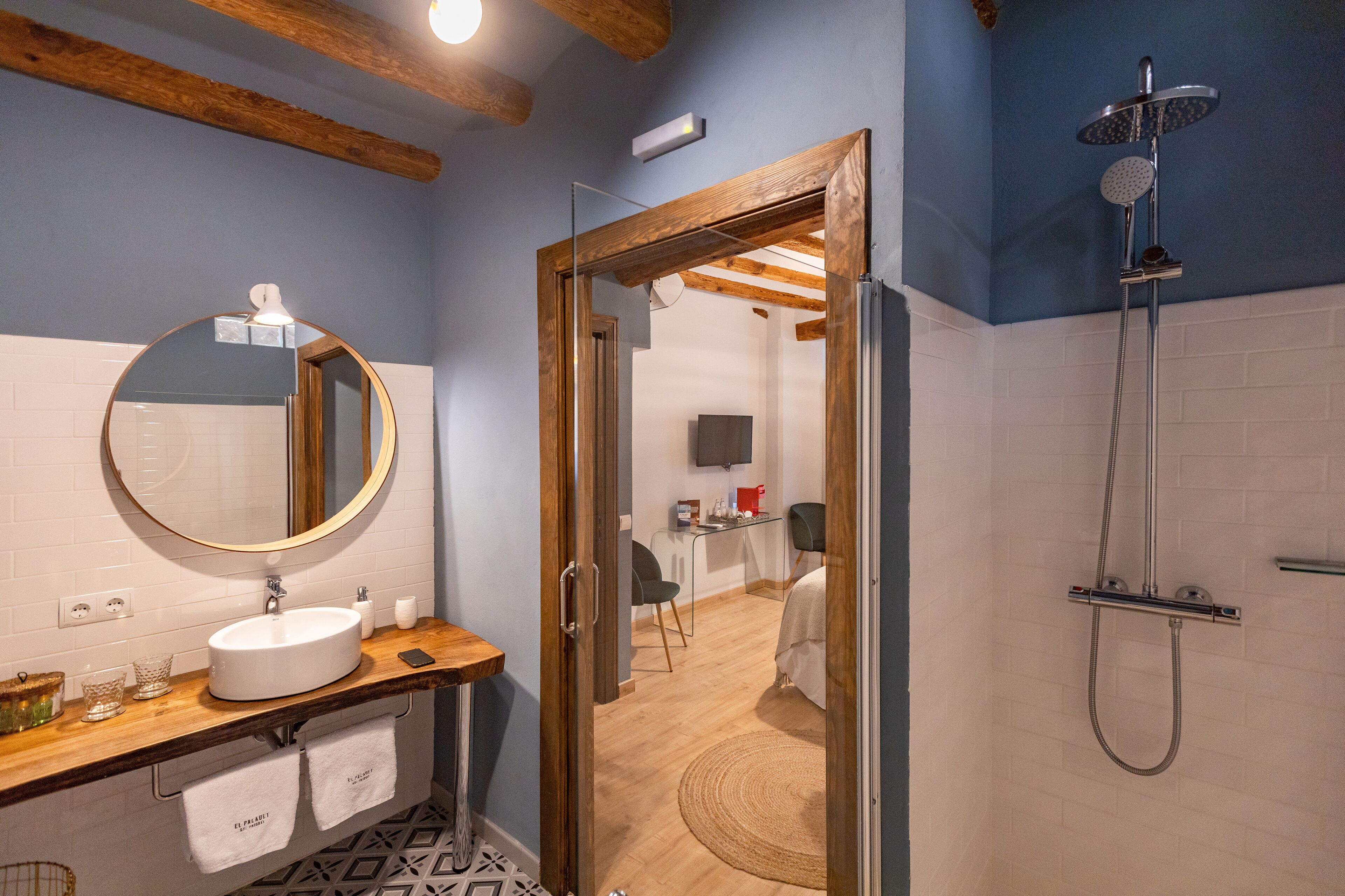 Un baño moderno con tocador de madera, un espejo redondo, ducha con cabezal de ducha manual y un televisor visible al fondo.