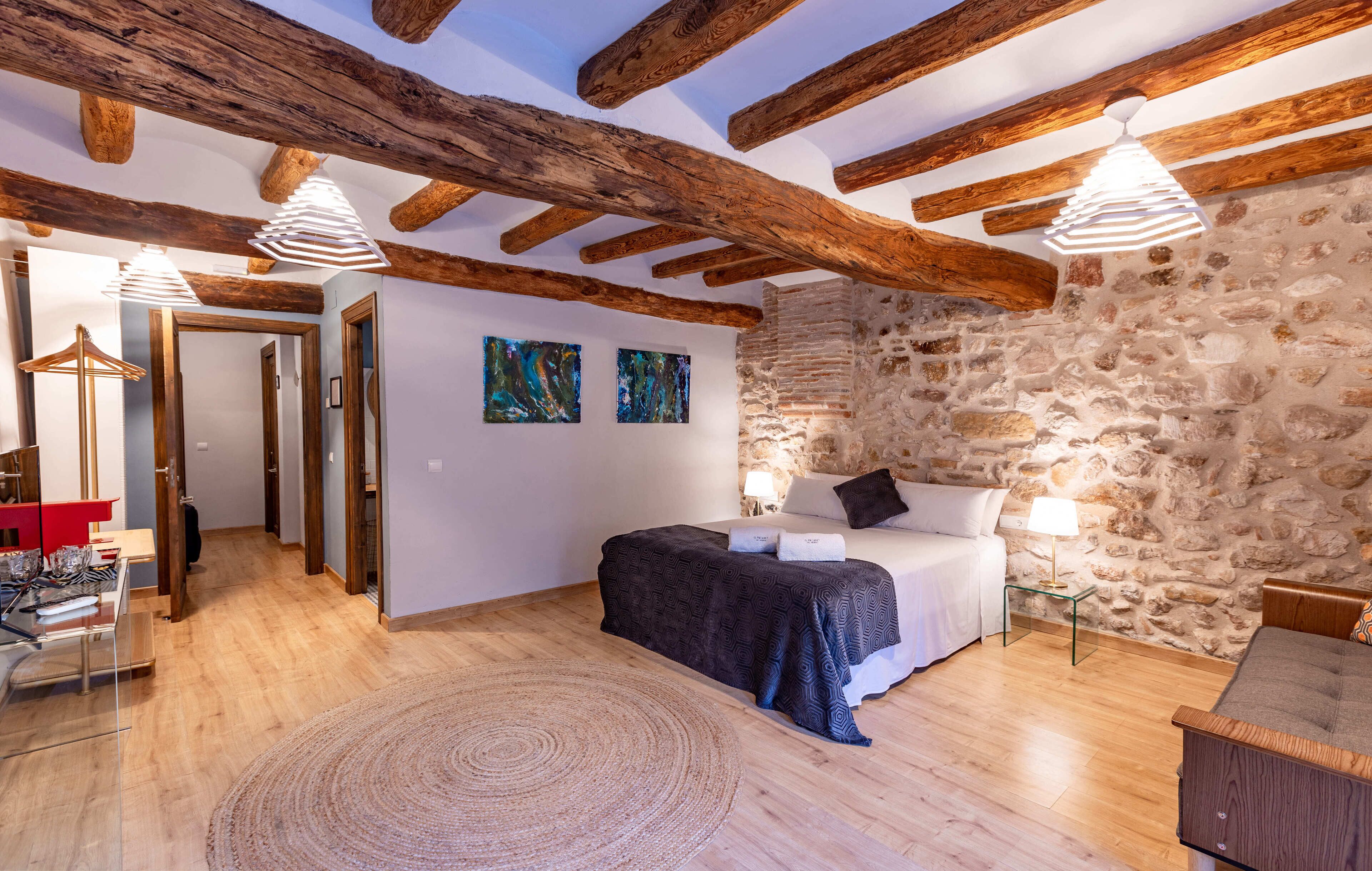 Un dormitorio con pared de piedra, vigas de madera, cama, mesita de noche y lámpara colgante.