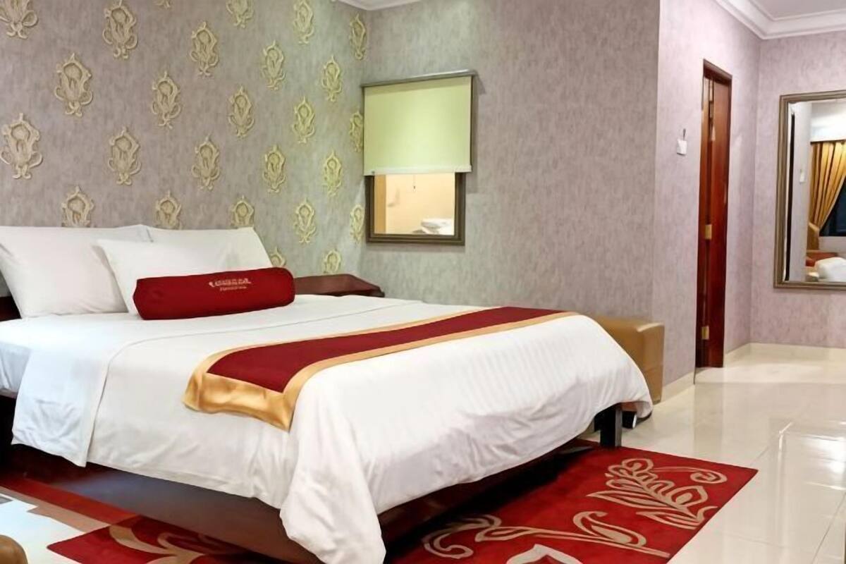 A hotel room with a large bed, a red throw blanket, a red pillow, a bedside table, a lamp, a mirror, and a red carpet.