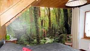 Een slaapkamer met een groot bed, een muurschildering van een bos, een houten plafond en een klein tafeltje met een groene beker.