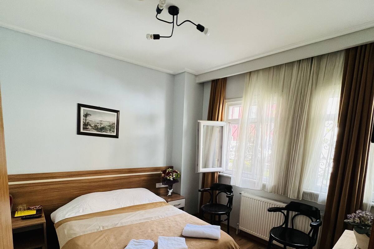 A hotel room with a large bed, a desk, a chair, a radiator, and a window with curtains.