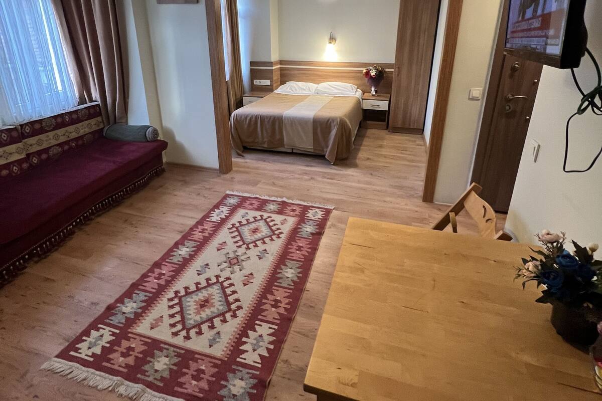 A hotel room with a bed, a wooden table, a red sofa, a patterned rug, and a ceiling light.