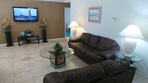 A living room with a brown leather sofa, a glass coffee table, two floor lamps, a TV on the wall, and a small plant on a console table. - Ly53774 - Indian Ridge - 4 Bed 2 Baths Villa (Kissimmee)