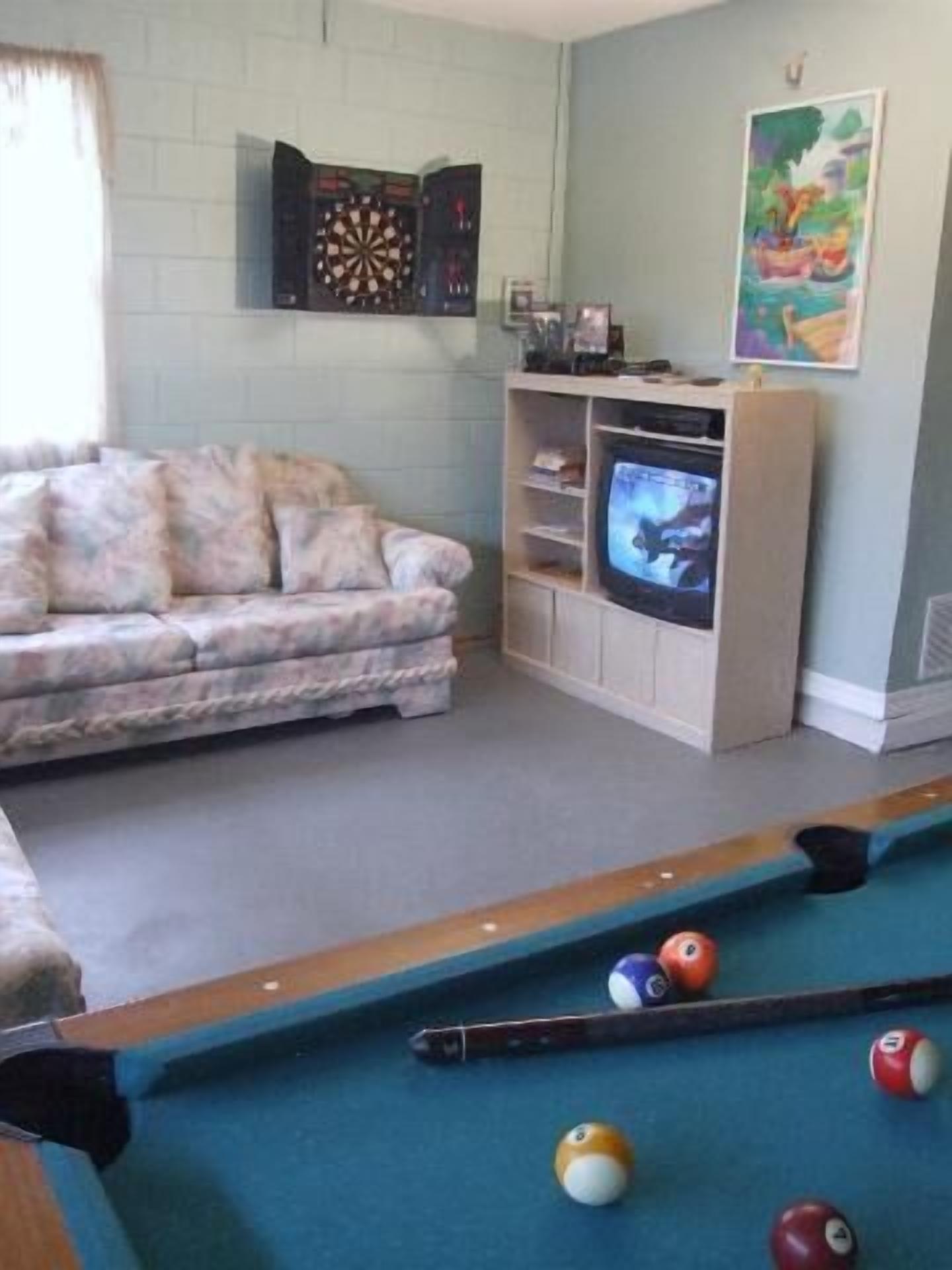 A living room with a pool table, a sofa, a television, and a dartboard.