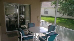 A covered patio with a round table and four chairs, a sliding glass door leading to a dining area, and a view of a house and lawn. - Ly53774 - Indian Ridge - 4 Bed 2 Baths Villa (Kissimmee)