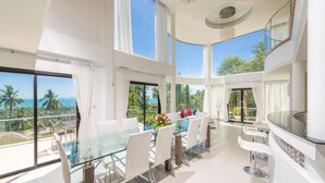 A spacious dining area with a glass table and white chairs, large windows offering a view of palm trees and the ocean, and a modern chandelier. - White Stone (Koh Samui)