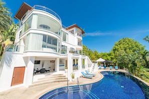 Una casa moderna de varios pisos con una gran piscina, mobiliario exterior y abundante vegetación.