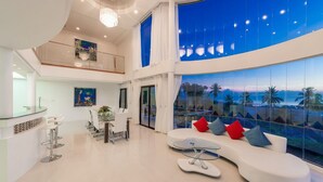 A modern interior with a curved white sofa, a dining area, and large windows offering a view of palm trees and a sunset. - White Stone (Koh Samui)
