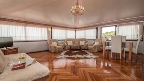 Un vaste salon doté d’un parquet en bois disposé en point de Hongrie, d’un lustre, de fauteuils en cuir, d’une table basse et d’un coin repas avec une table et des chaises blanches.