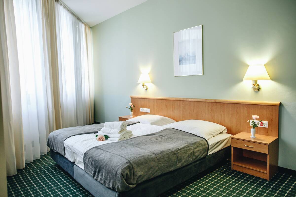 A hotel room with a bed, two bedside tables, a nightstand, a lamp, a framed picture on the wall, and a window with curtains.