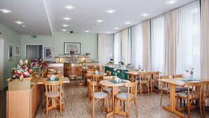 Un comedor de hotel con mesas y sillas de madera, una cocina al fondo y amplias ventanas con cortinas translúcidas.