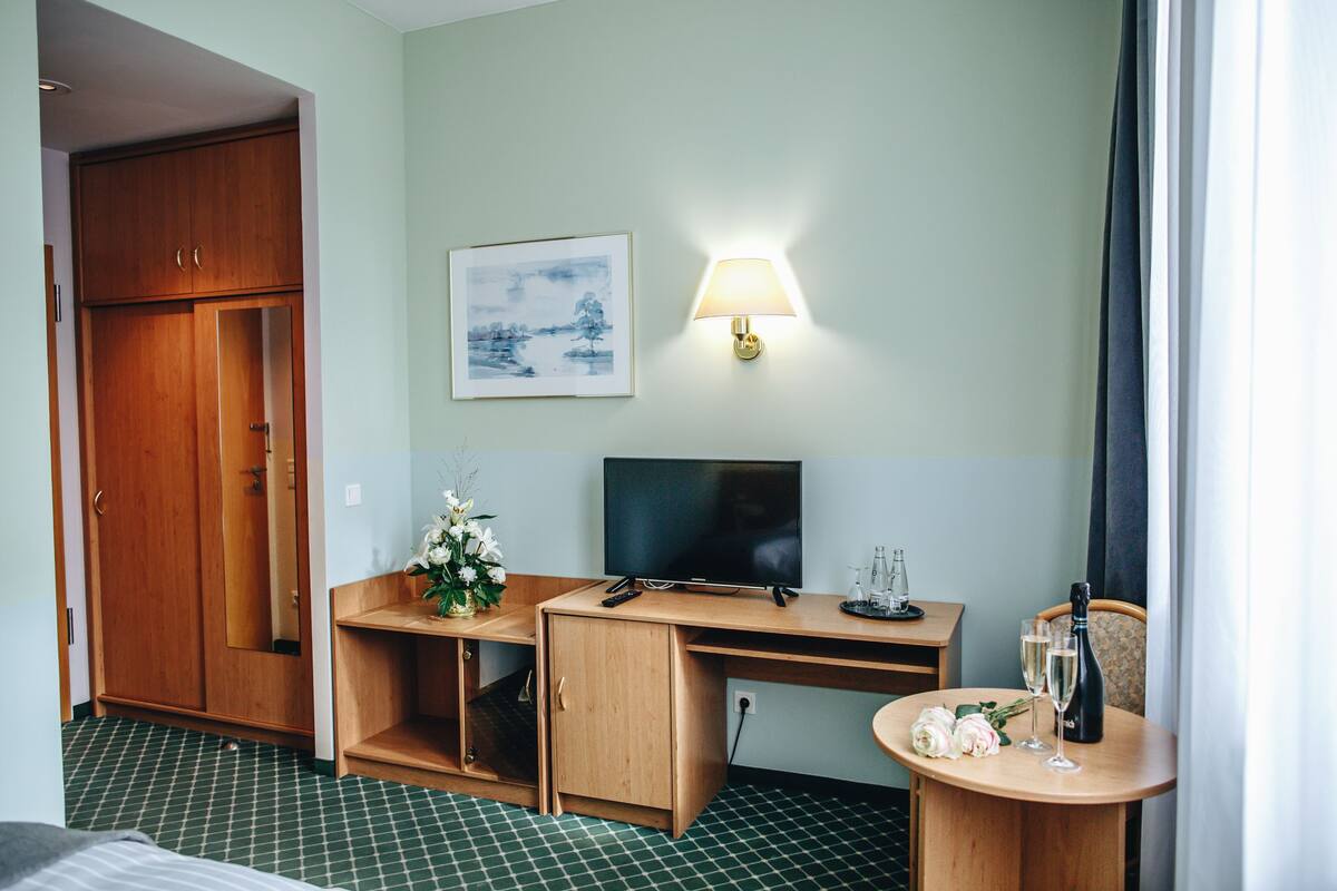 A hotel room with a wooden desk, a television, a bedside table with a bottle and glasses, a mirror, and a wardrobe.