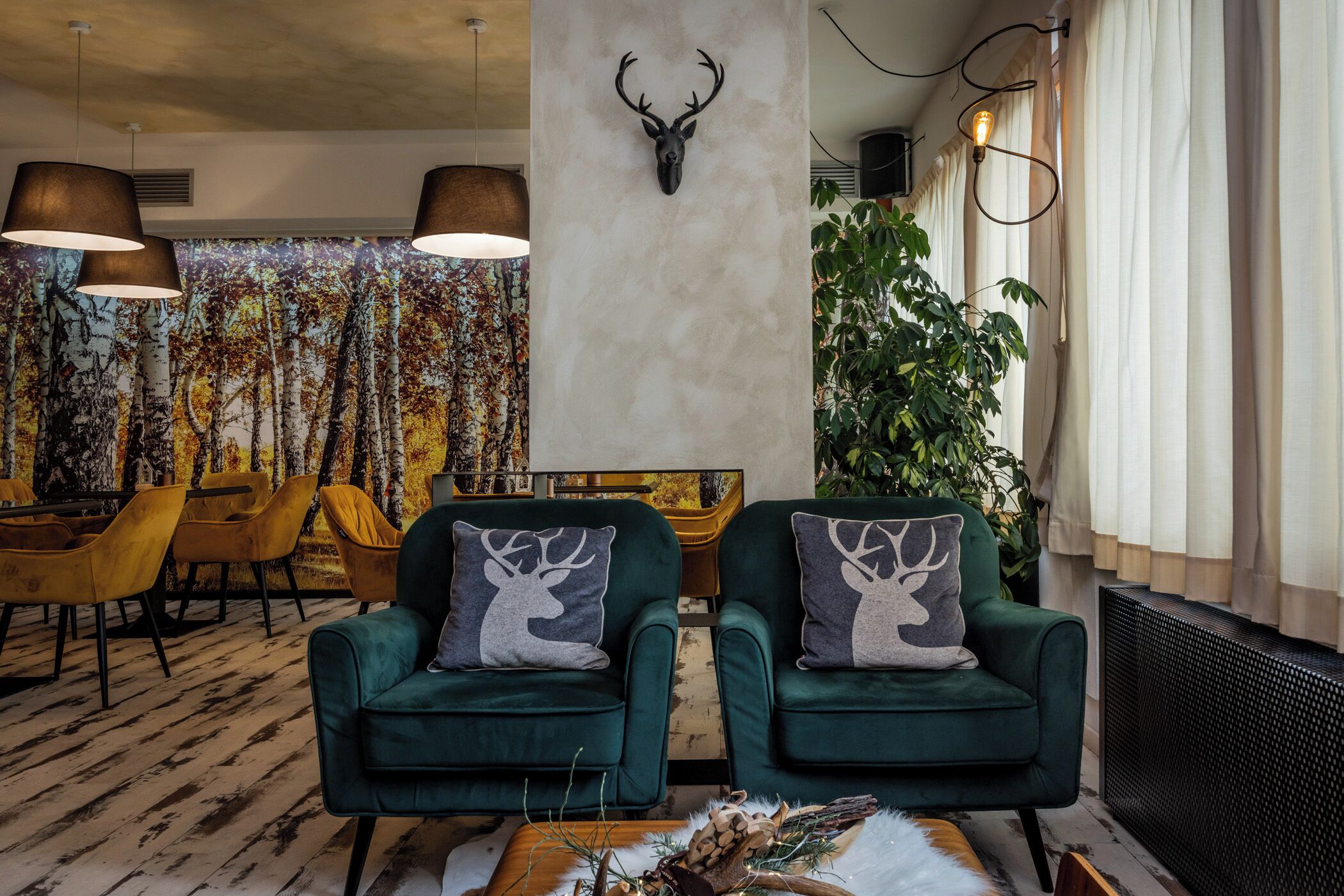 A room with two green armchairs, a deer-themed pillow, a deer head mounted on the wall, a large painting of trees, and a plant.