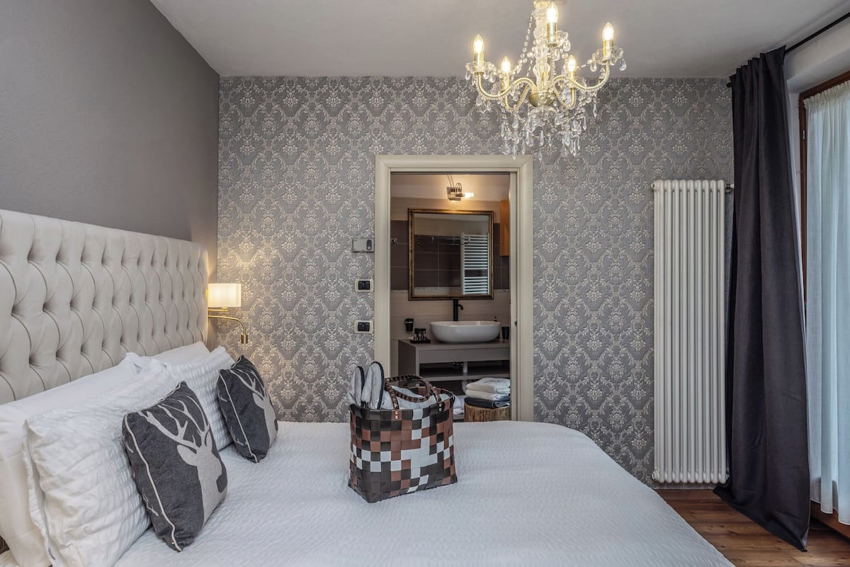 Ein Schlafzimmer mit einer gepolsterten Kopfteile, einem großen Bett, einer Deckenleuchte, einem Badezimmer mit Waschbecken und Spiegel sowie einem Heizkörper.