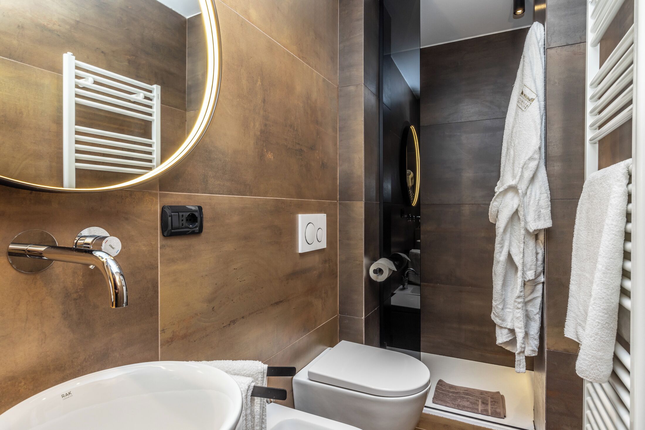 Un baño moderno con lavamanos blanco, inodoro, toallero y espejo con luz circular.