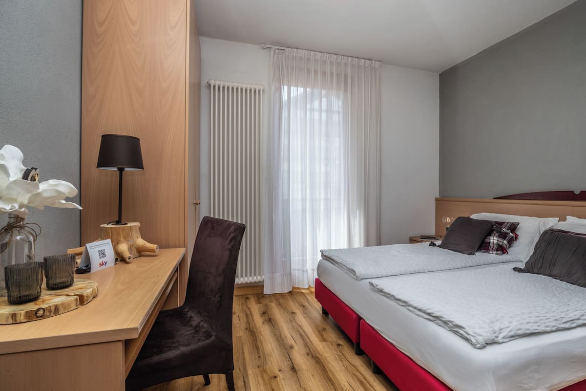 Ein Hotelzimmer mit einem Holztisch, einem Sessel, einem Bett mit roten Akzenten und einem Fenster mit durchscheinenden Vorhängen.