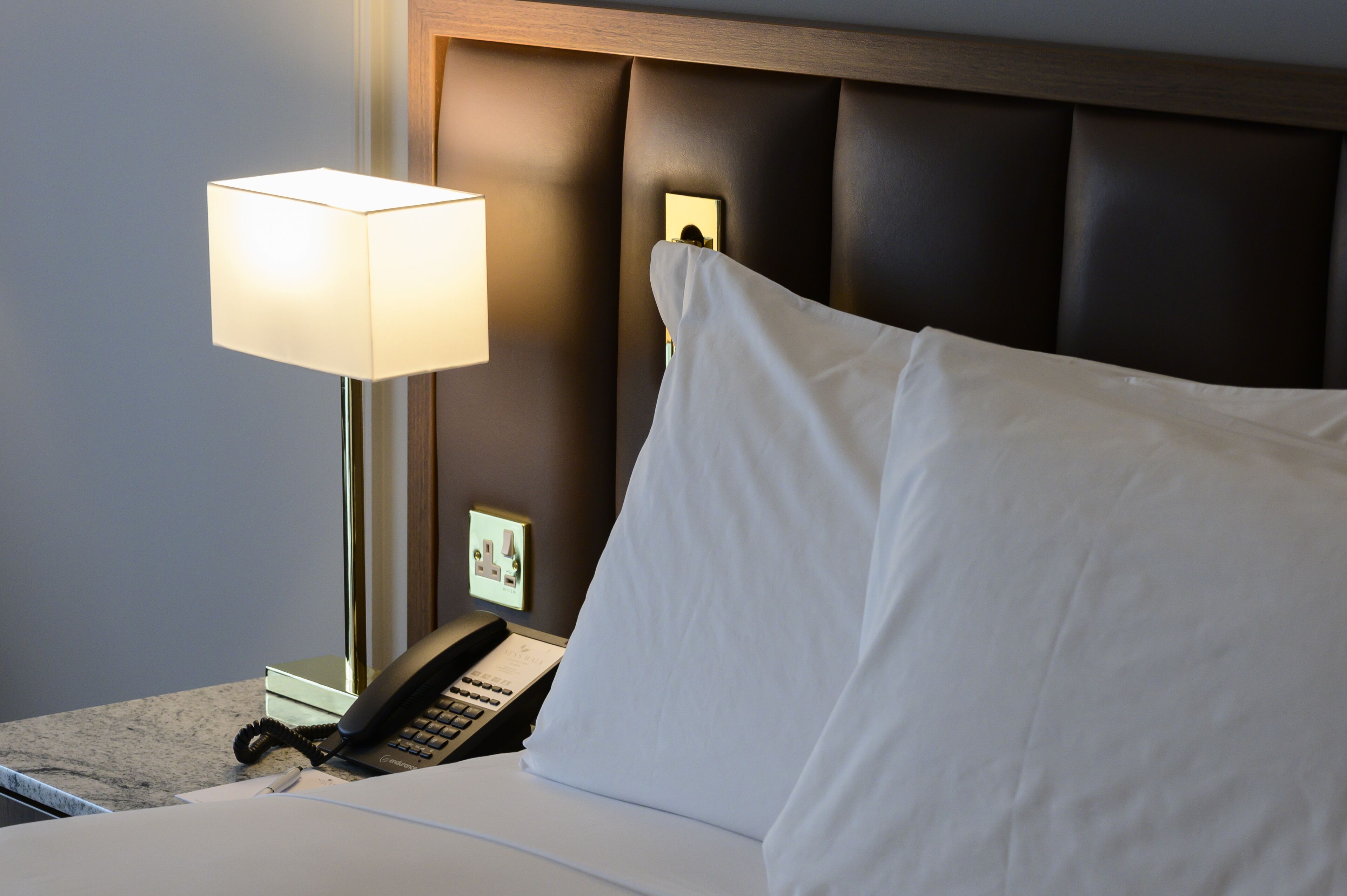 A hotel room with a bed, a bedside lamp, a telephone, and a small picture frame on the nightstand.