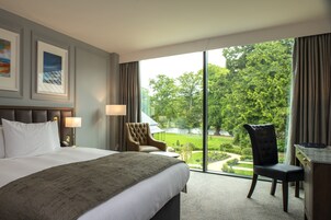 Habitación de hotel con una cama grande, dos sillas, un escritorio y vistas a un jardín a través de un amplio ventanal.