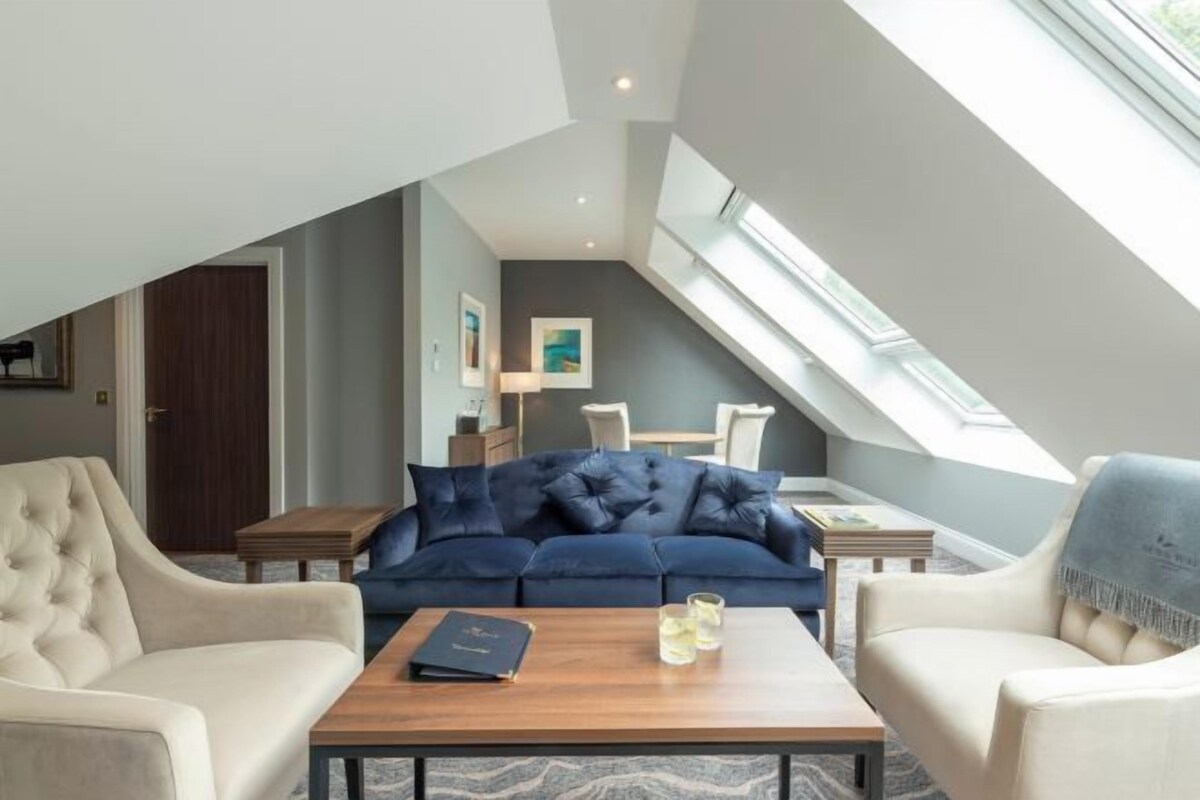 A modern loft bedroom with a sloped ceiling, a dark blue sofa, two armchairs, a wooden coffee table, and a skylight.