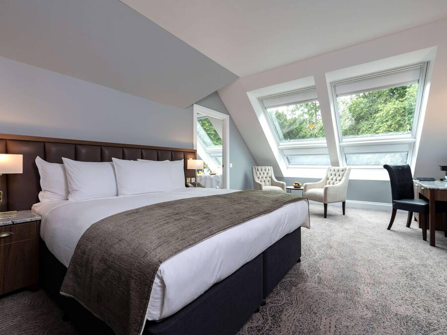A hotel room with a large bed, a desk, and a chair. There are two windows providing natural light.