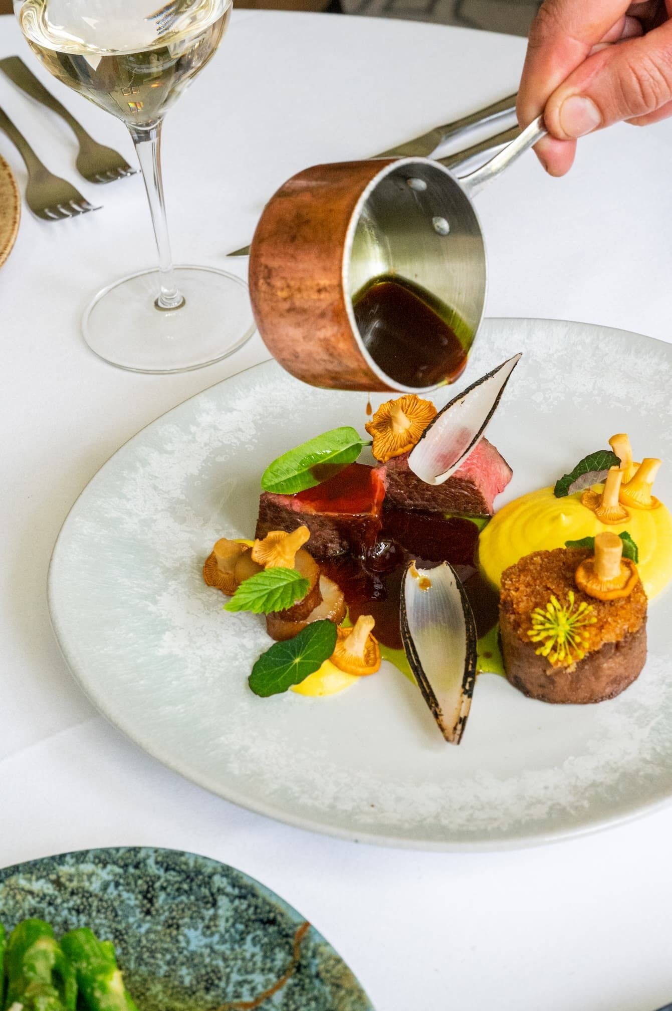 A person pouring a dark liquid from a copper cup onto a plate with a variety of food items, including a piece of meat and mushrooms.