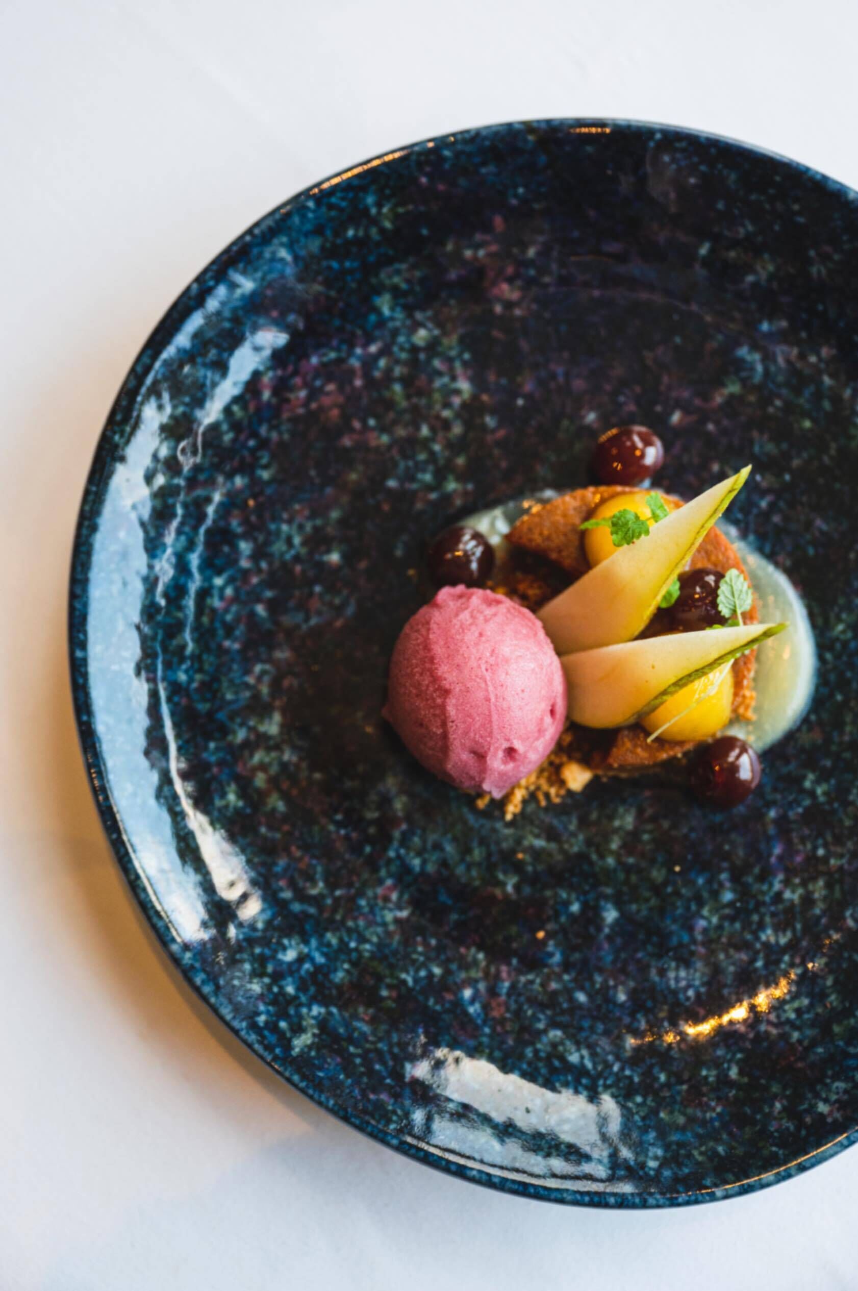 En dessert på en mørk, stribet tallerken med en kugle lyserød is, skiver af pære og kirsebær.