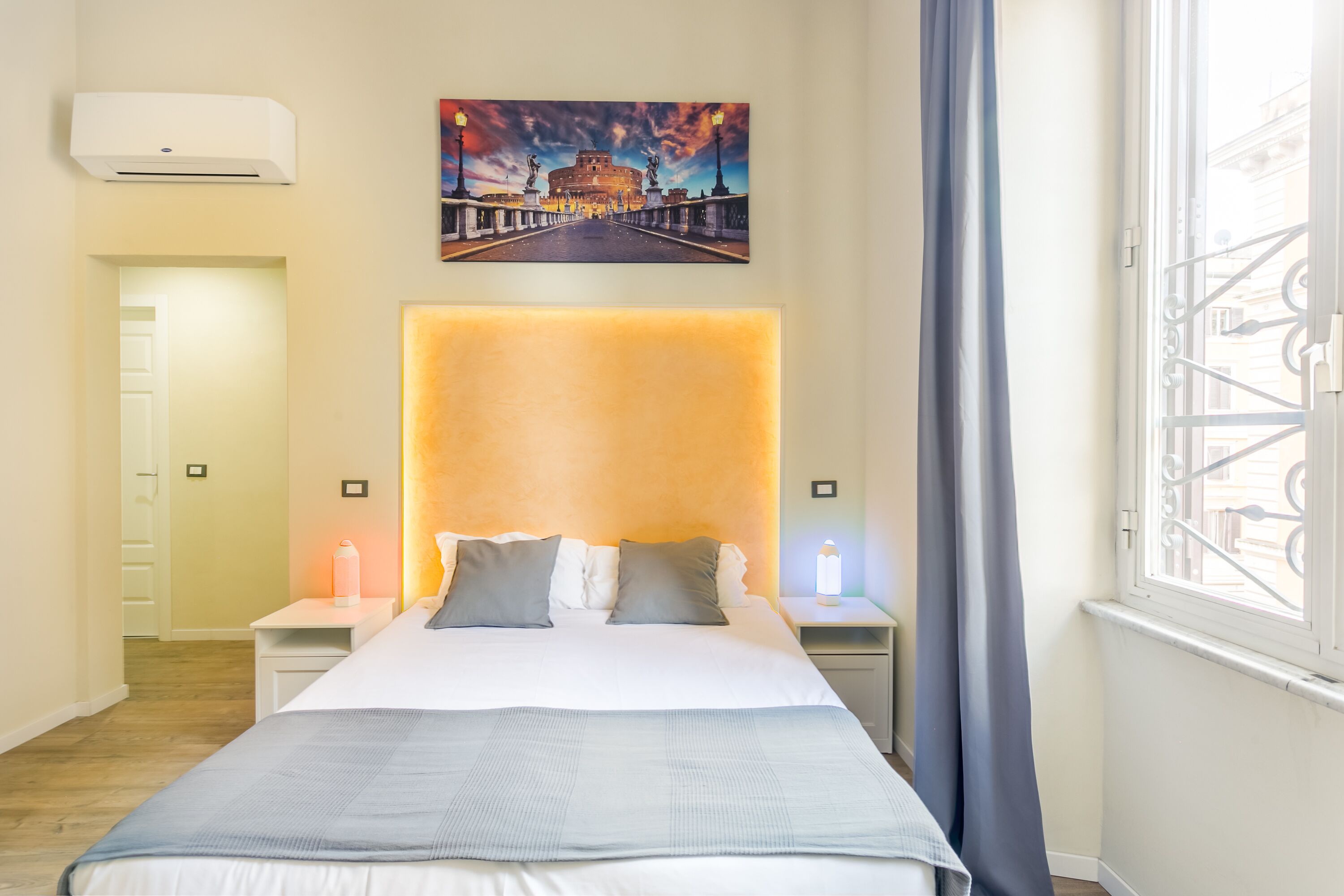 A hotel room with a large bed, a headboard with a light panel, a painting of a bridge, and a window with metal bars.