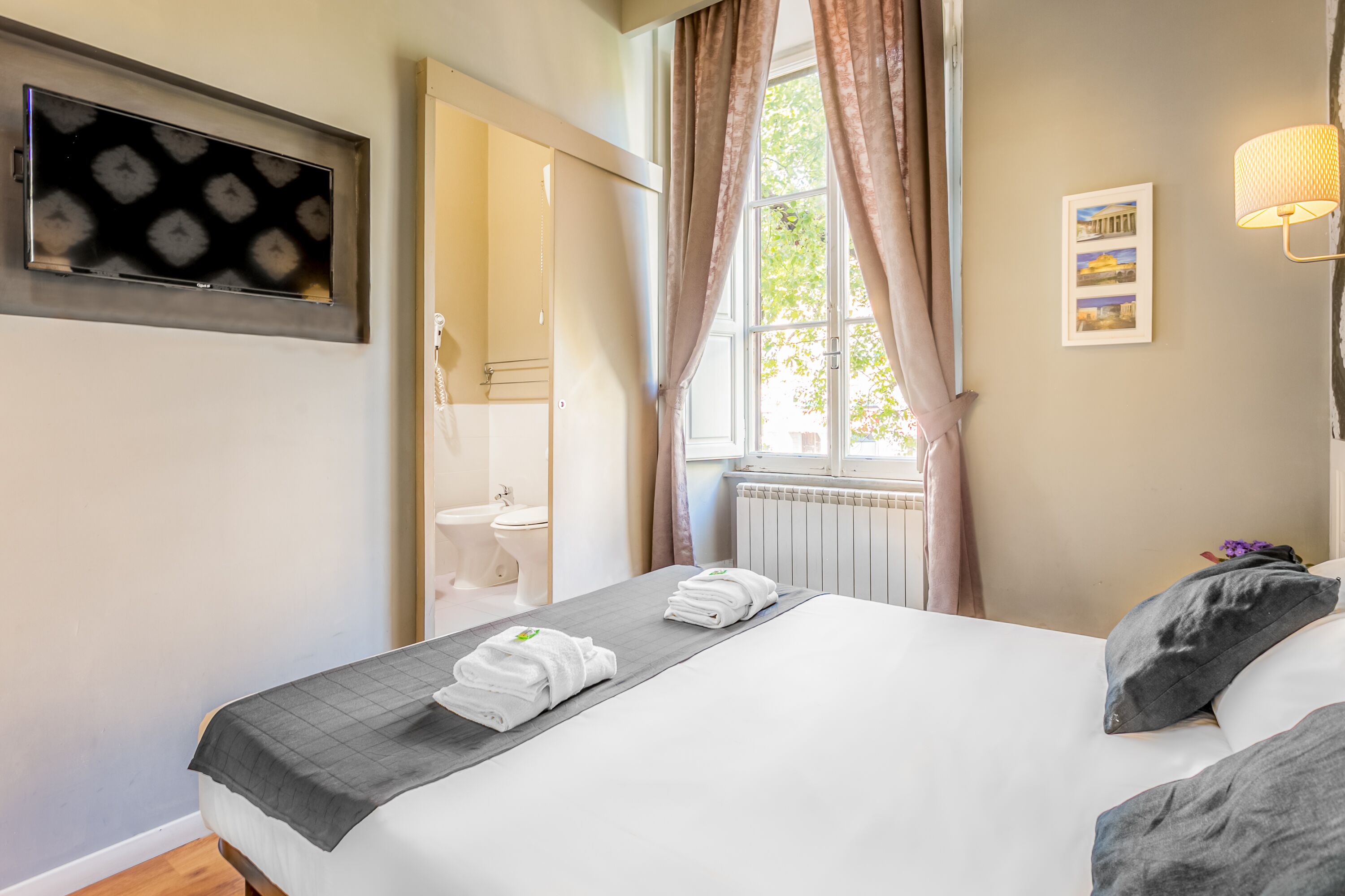 A hotel room with a bed, a TV mounted on the wall, a window with curtains, and a radiator.