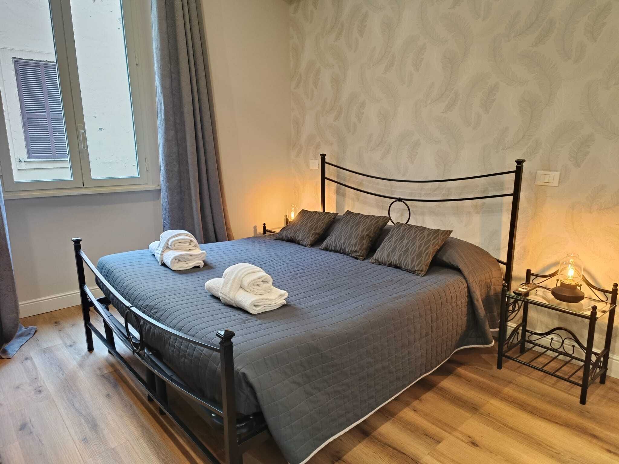Una cama bien hecha con estructura metálica, ropa de cama gris y dos toallas enrolladas a un lado.