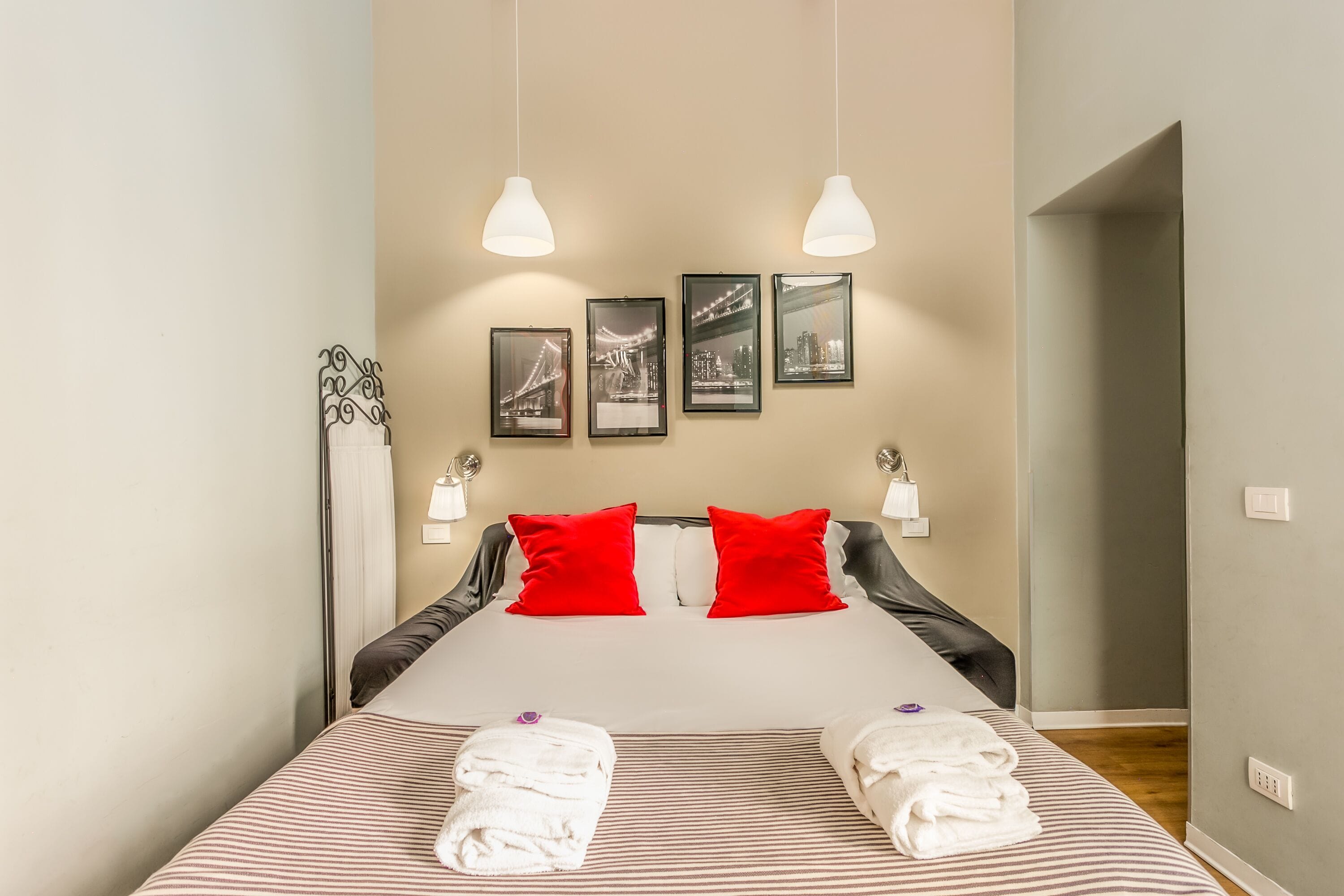 A neatly made bed with striped bedding, two red pillows, and two folded towels. There are three framed pictures on the wall above the bed.