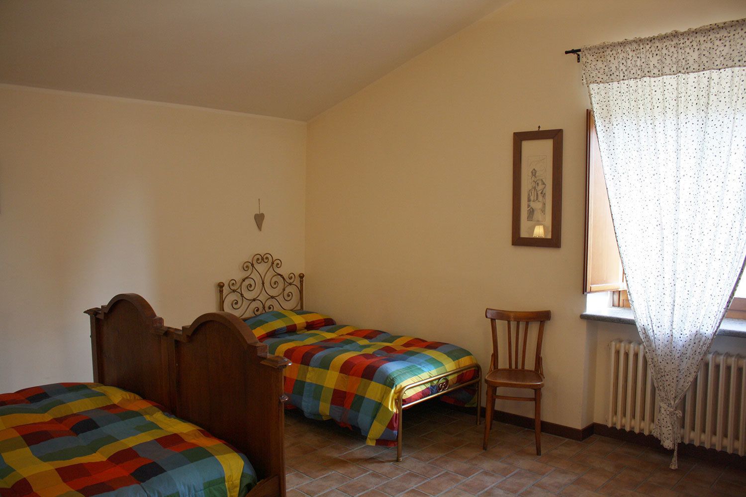A bedroom with two single beds, a wooden chair, a radiator, and a window with a curtain.