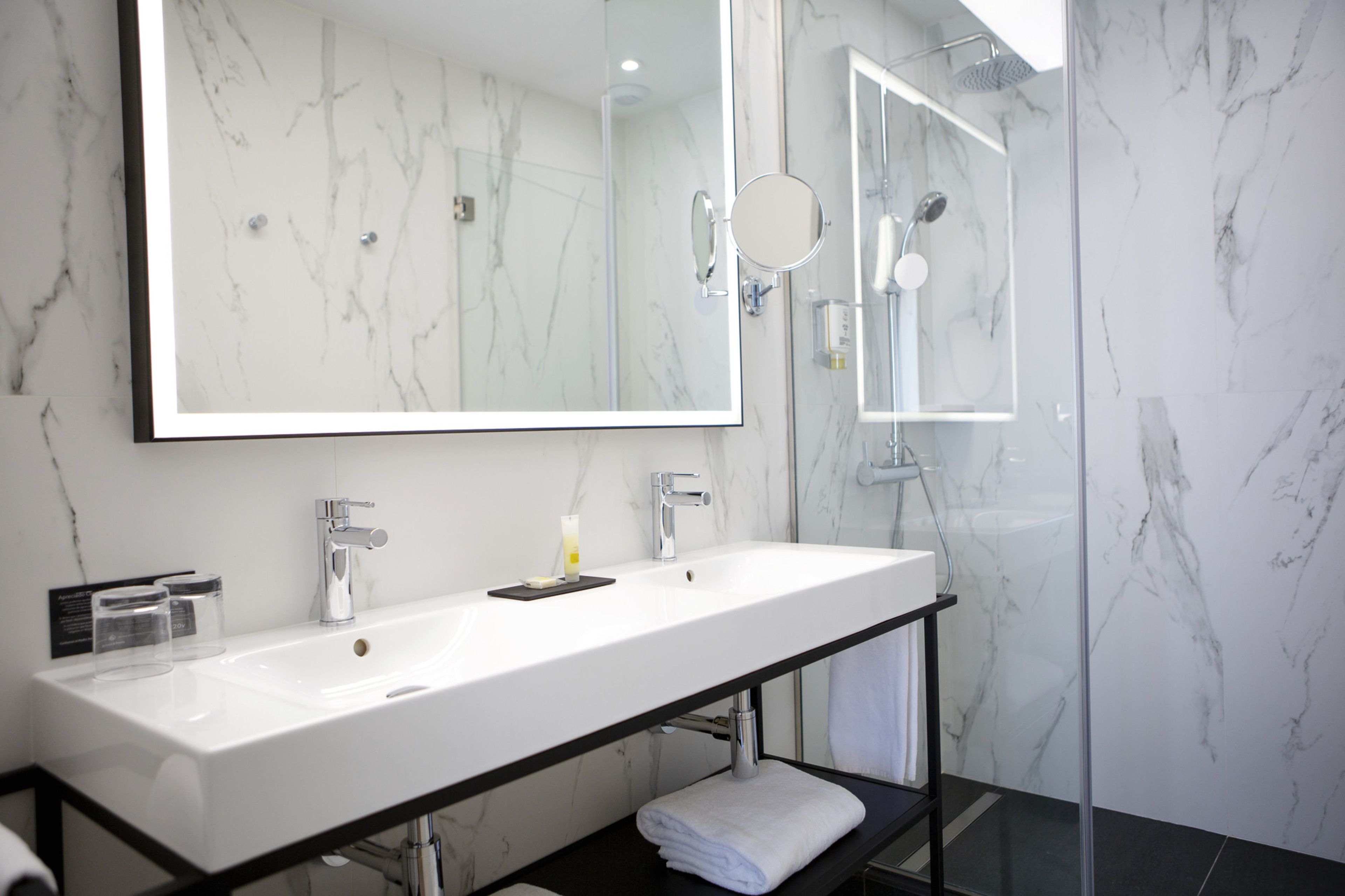 Une salle de bain moderne avec un grand miroir, deux lavabos, une douche et un mur en marbre.