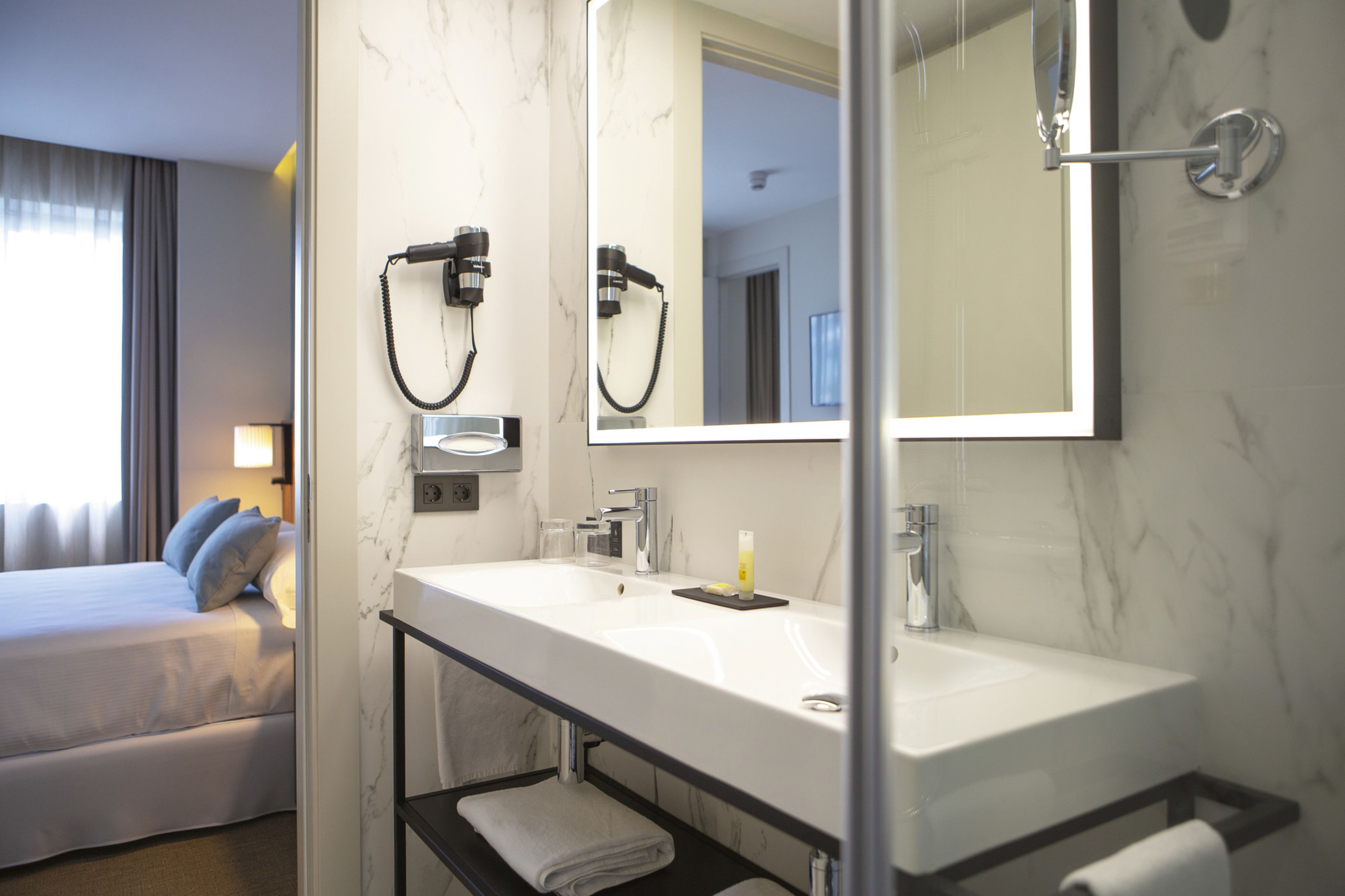 A hotel bathroom with a large mirror, a sink, and a shower. There is a bed with a view of the outside.