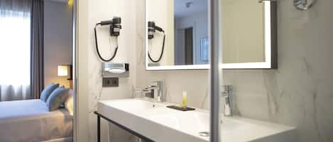 A hotel bathroom with a large mirror, a sink, and a shower. There is a bed with a view of the outside.