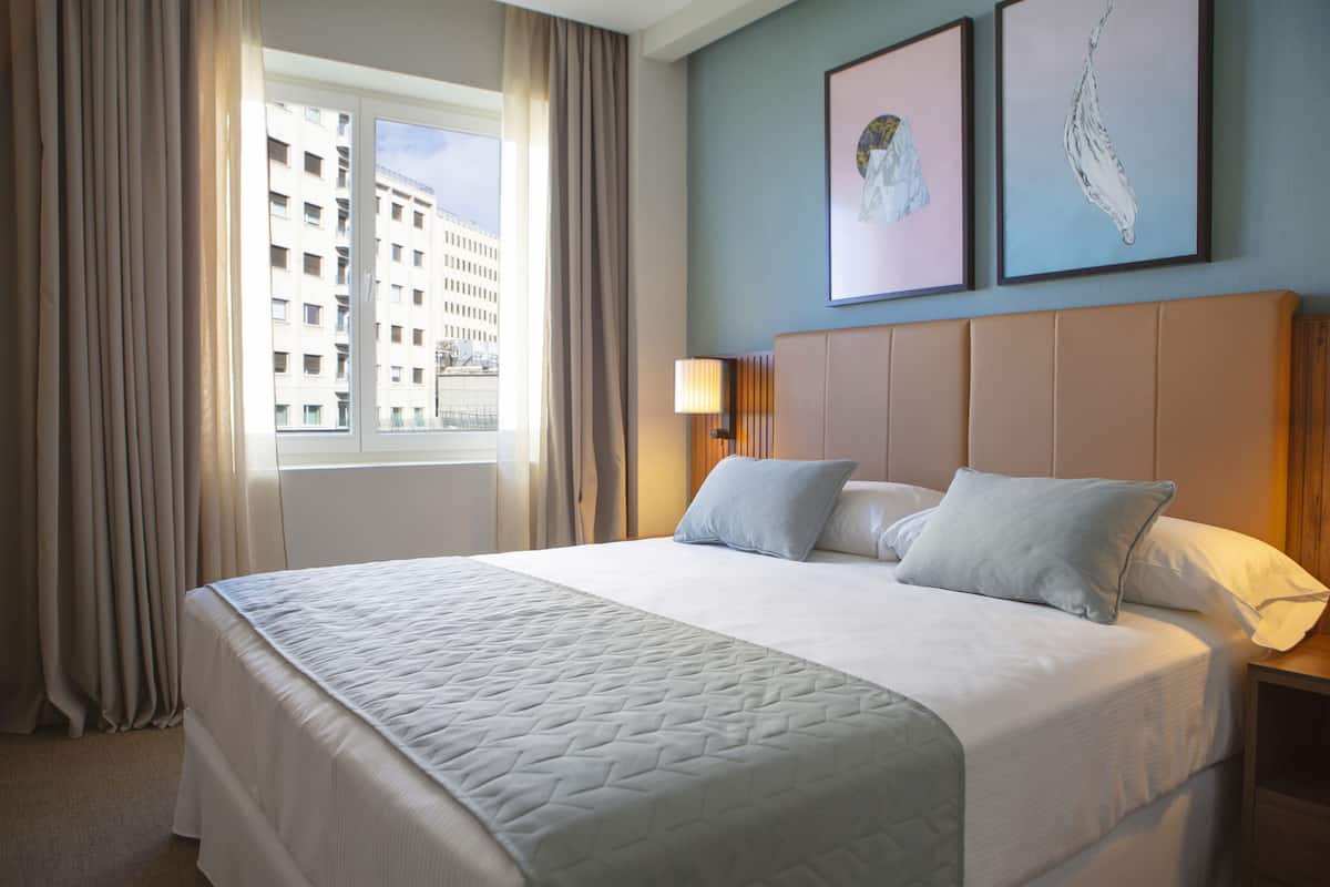 A hotel room with a large bed, a window with curtains, a headboard, and two framed artworks on the wall.