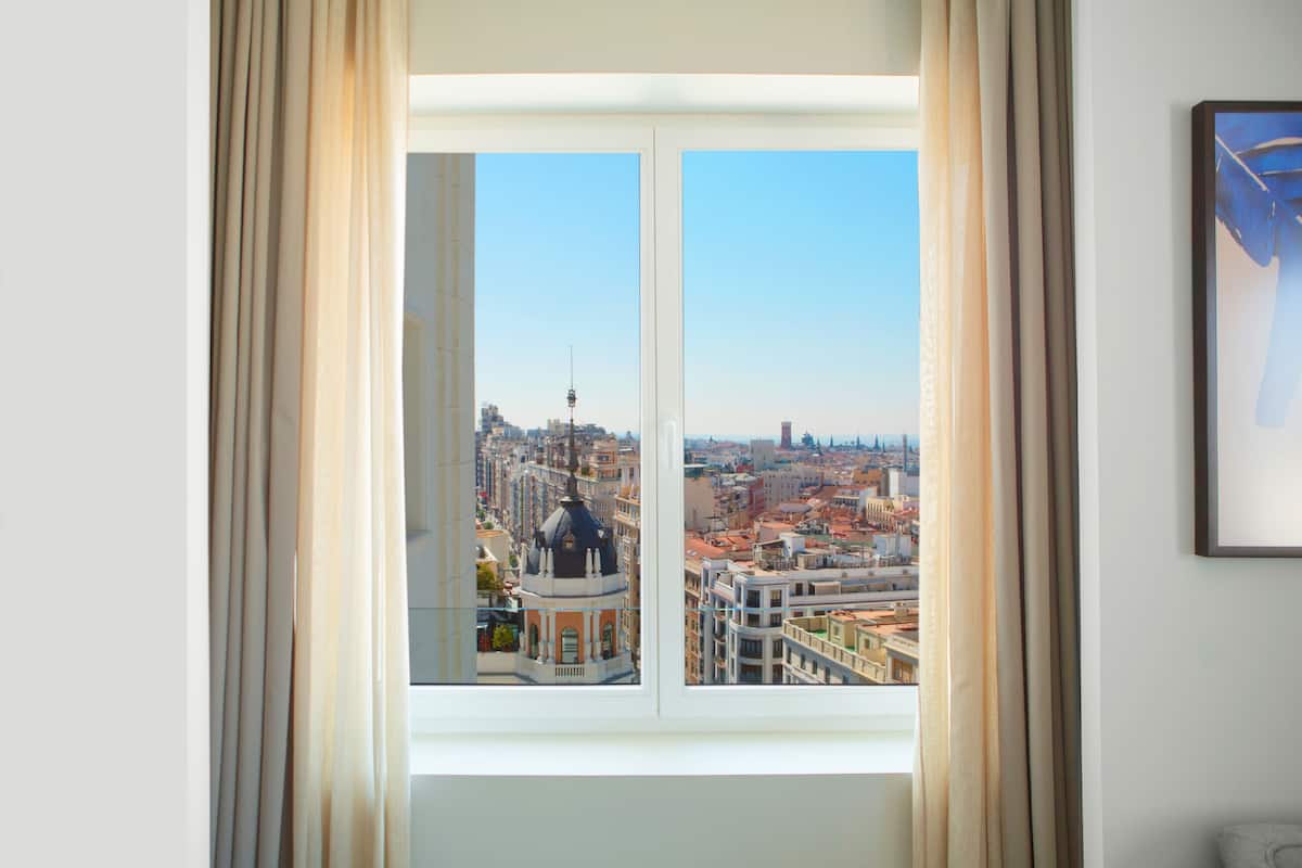 A room with a large window offering a cityscape view, curtains, and a framed picture on the wall.