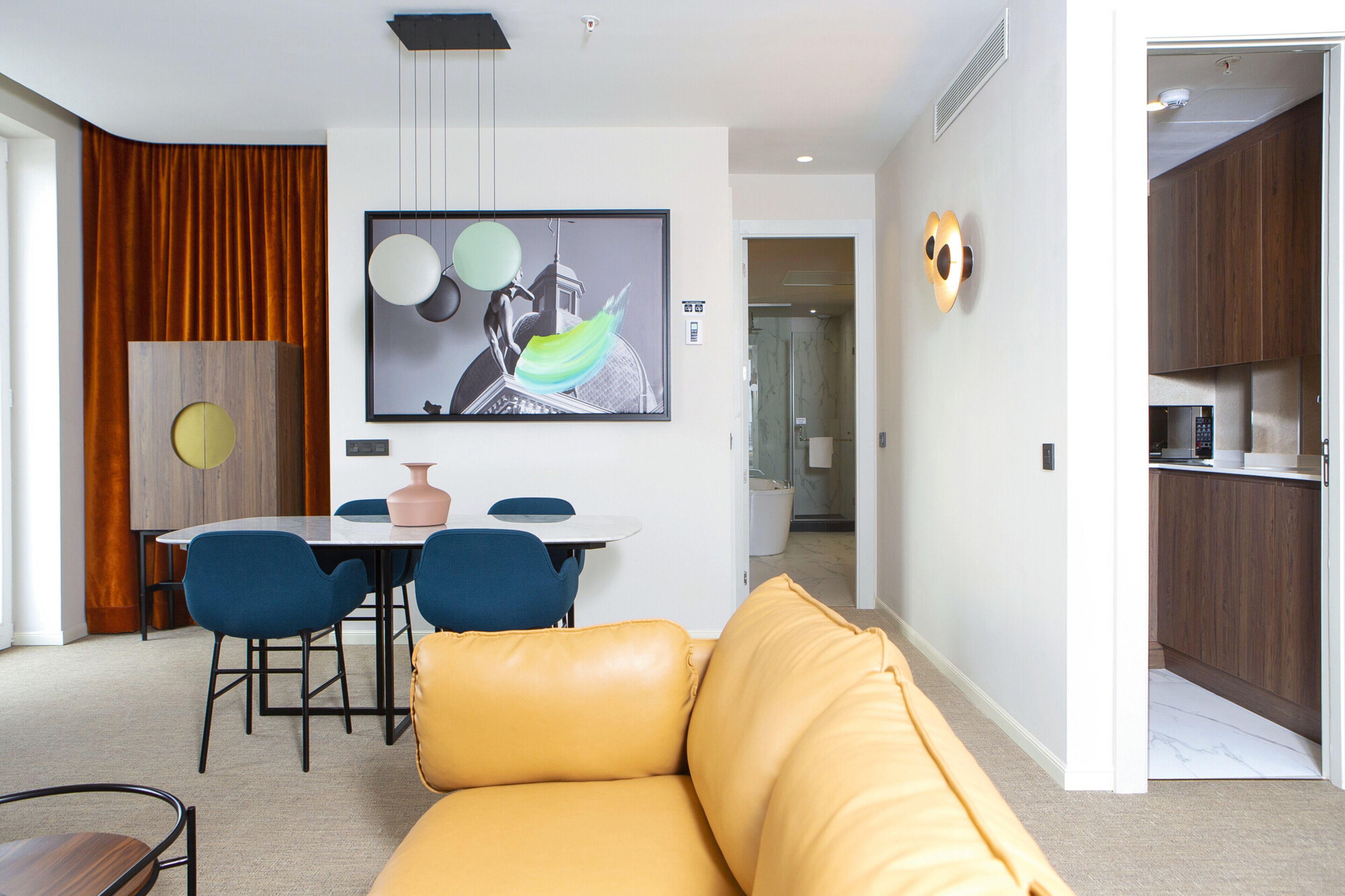 Una sala moderna con un sofá amarillo, una zona de comedor con sillas azules y una cocina visible a través de una puerta abierta.