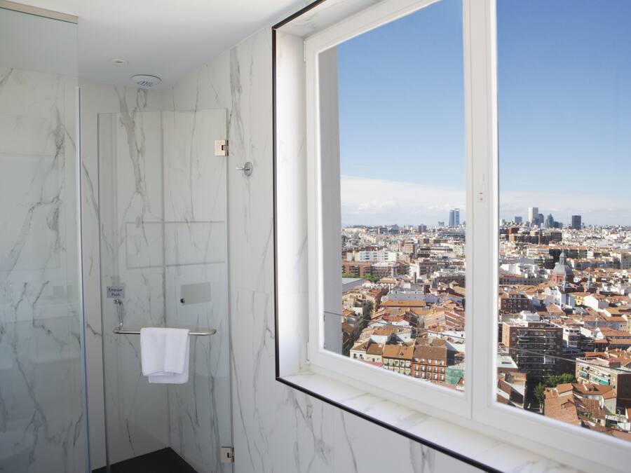 Un baño con mampara de vidrio y ventana con vista a la ciudad.