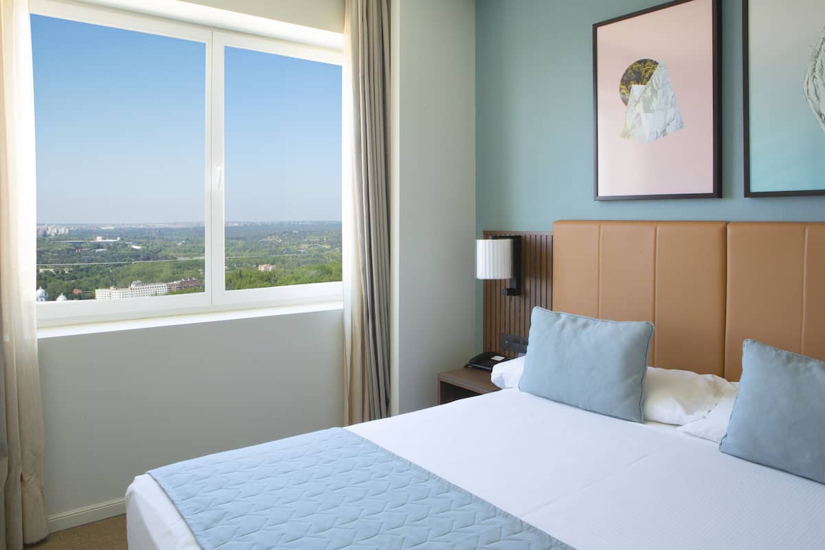 A hotel room with a large bed, a view of the city, a nightstand, and a wall with two framed pictures.