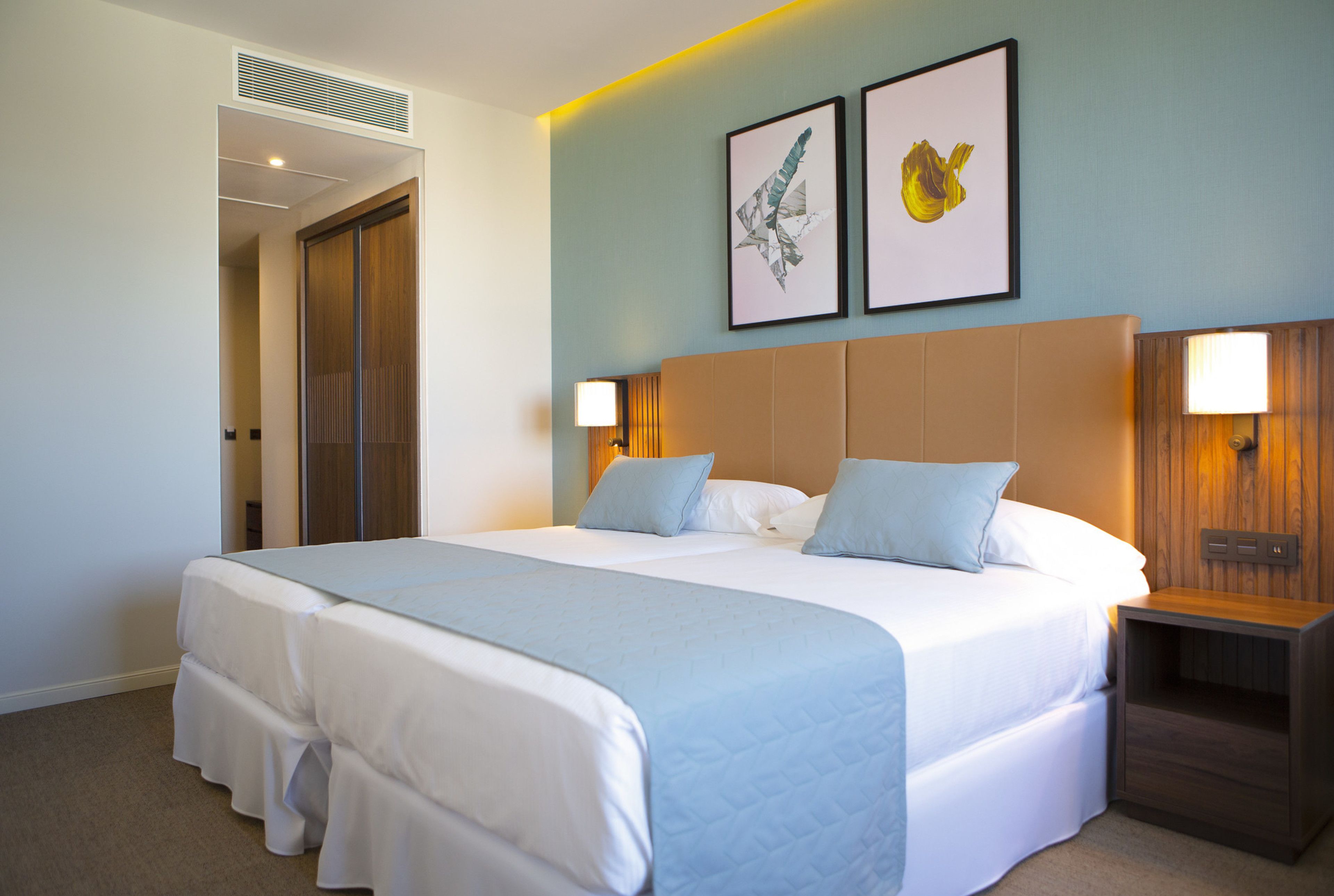 A hotel room with a large bed, two bedside tables with lamps, a wooden headboard, and two framed artworks on the wall.