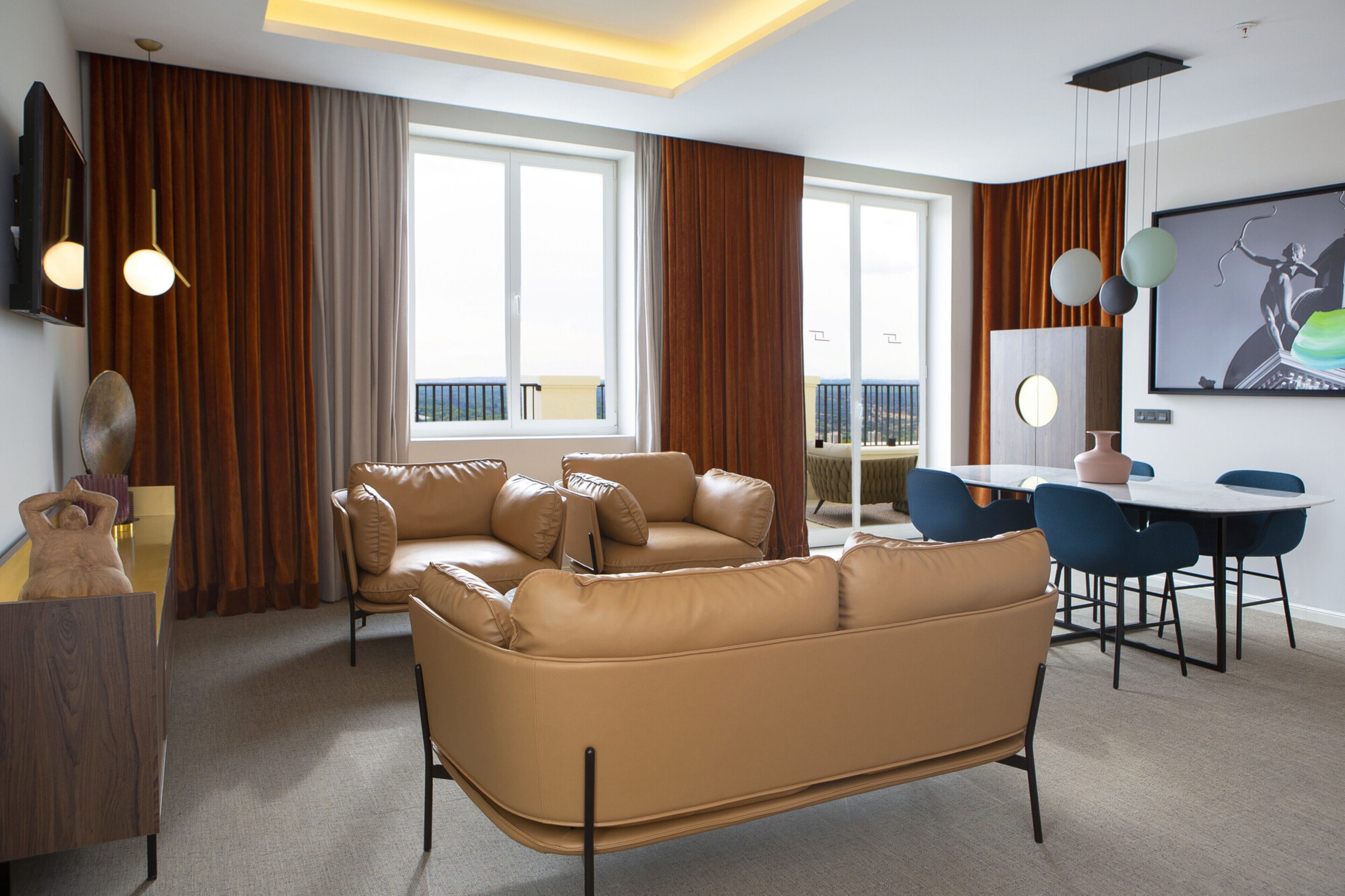 A modern living room with a brown leather sofa, a dining table with chairs, and large windows with curtains.