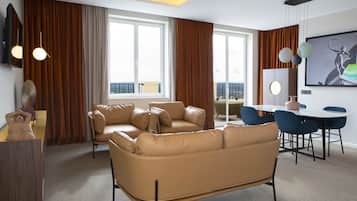 A modern living room with a brown leather sofa, a dining table with chairs, and large windows with curtains.