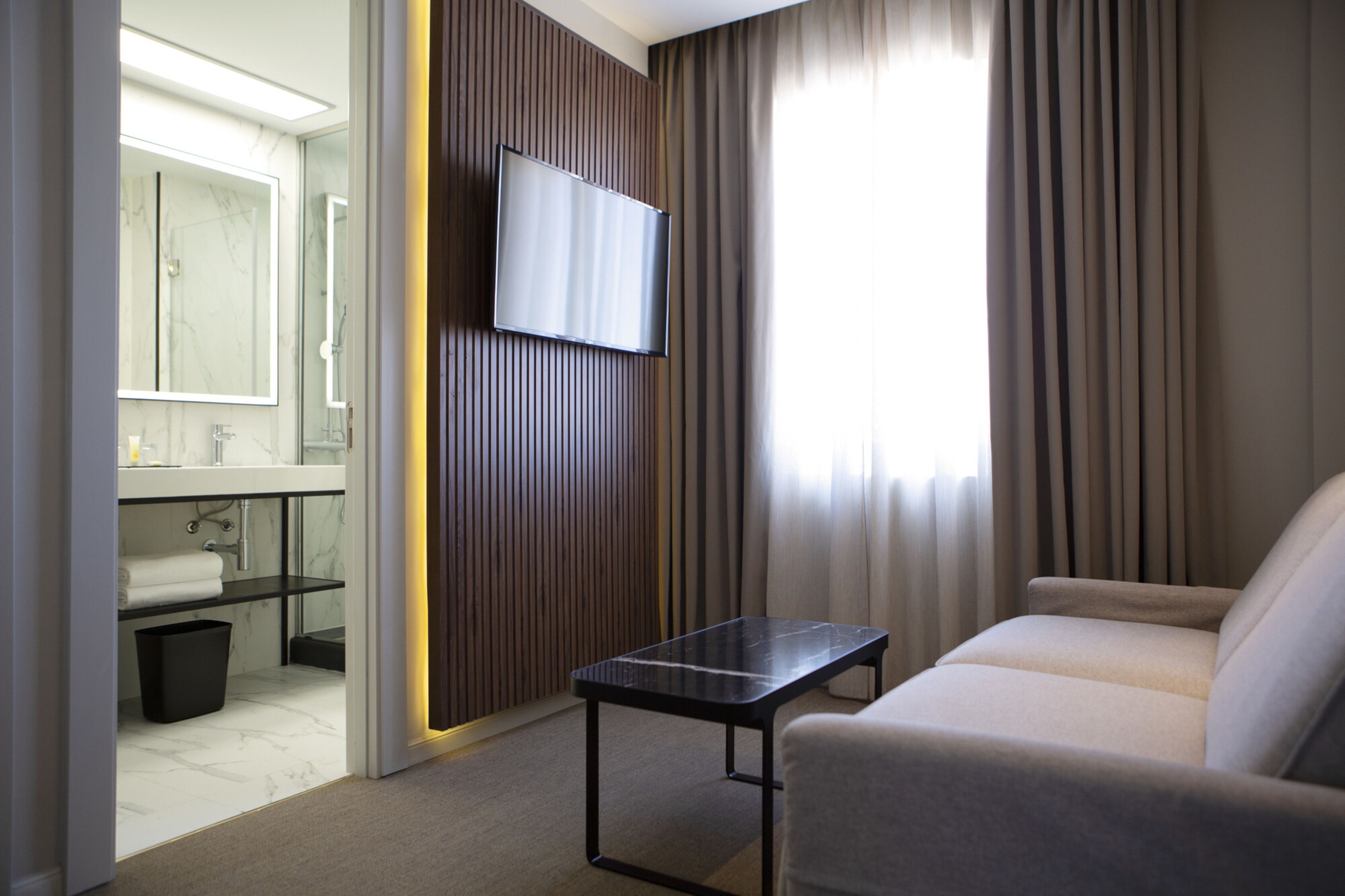 Une chambre d’hôtel avec un canapé, un téléviseur fixé sur un panneau en bois, une salle de bain avec un miroir et un lavabo, et une fenêtre avec des rideaux.