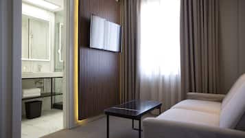 Une chambre d’hôtel avec un canapé, un téléviseur fixé sur un panneau en bois, une salle de bain avec un miroir et un lavabo, et une fenêtre avec des rideaux.