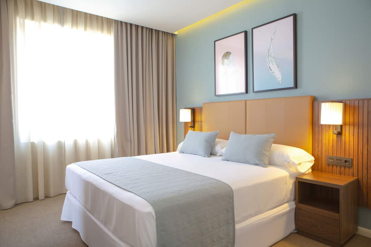 A hotel room with a large bed, two bedside lamps, a nightstand, and a window with curtains.