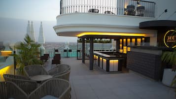A rooftop bar with wicker chairs and a bar counter, overlooking a cityscape with a prominent tower.