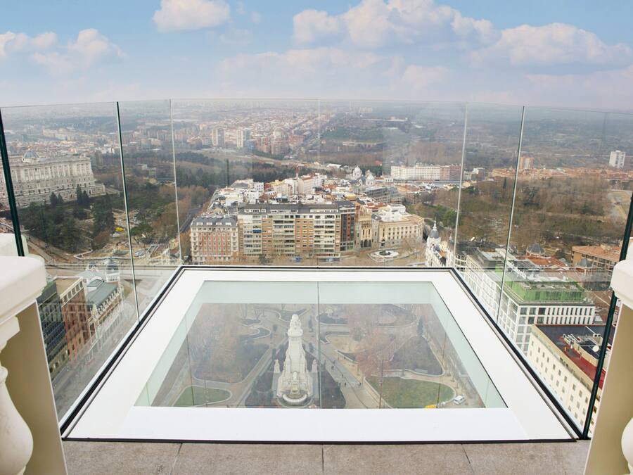 Um edifício alto com piso de vidro, oferecendo vista de um horizonte urbano com diversos edifícios e uma grande estátua ao centro.