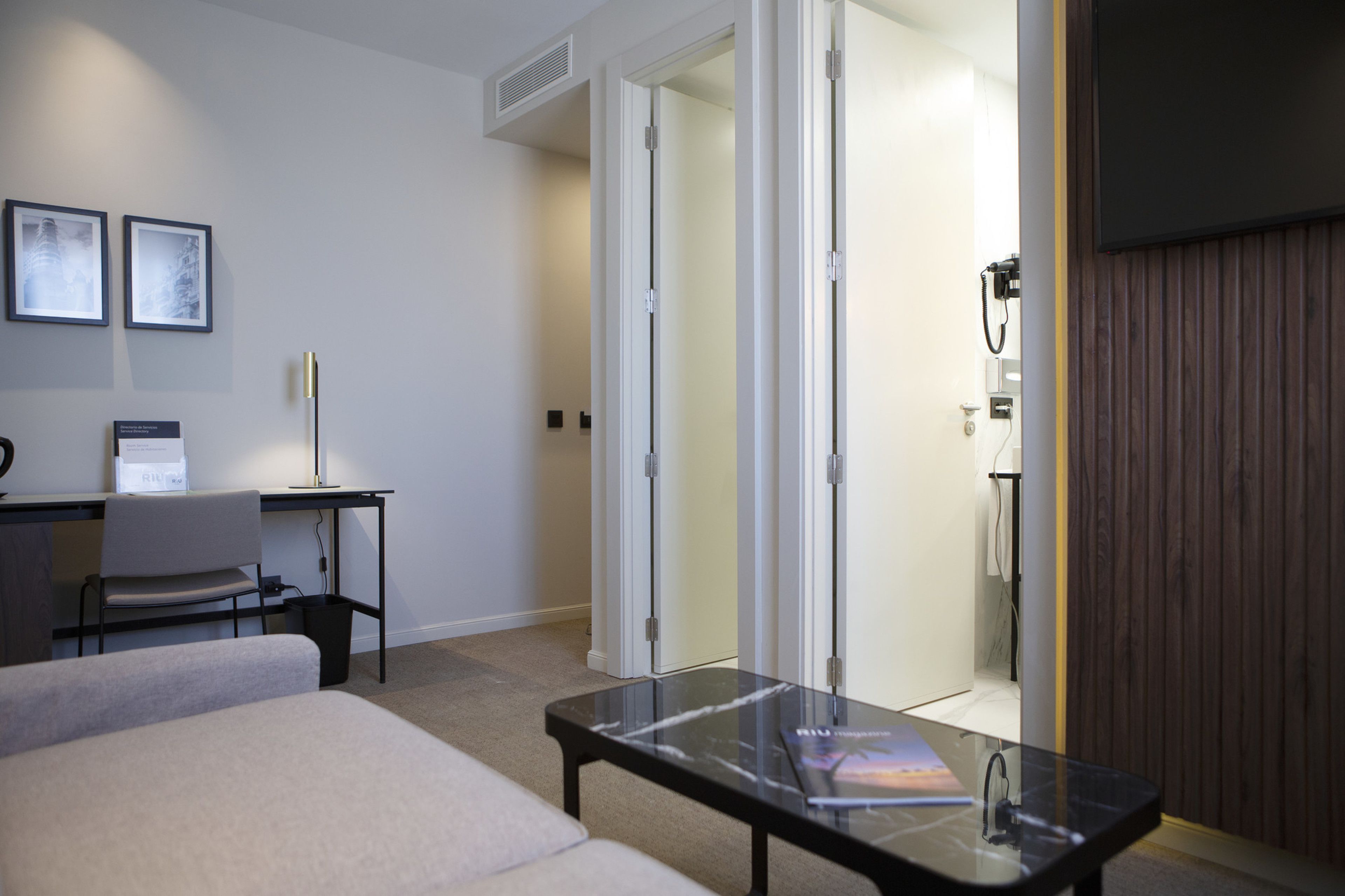 A modern hotel room with a bed, a desk, a chair, a glass table, and a wooden panel wall.