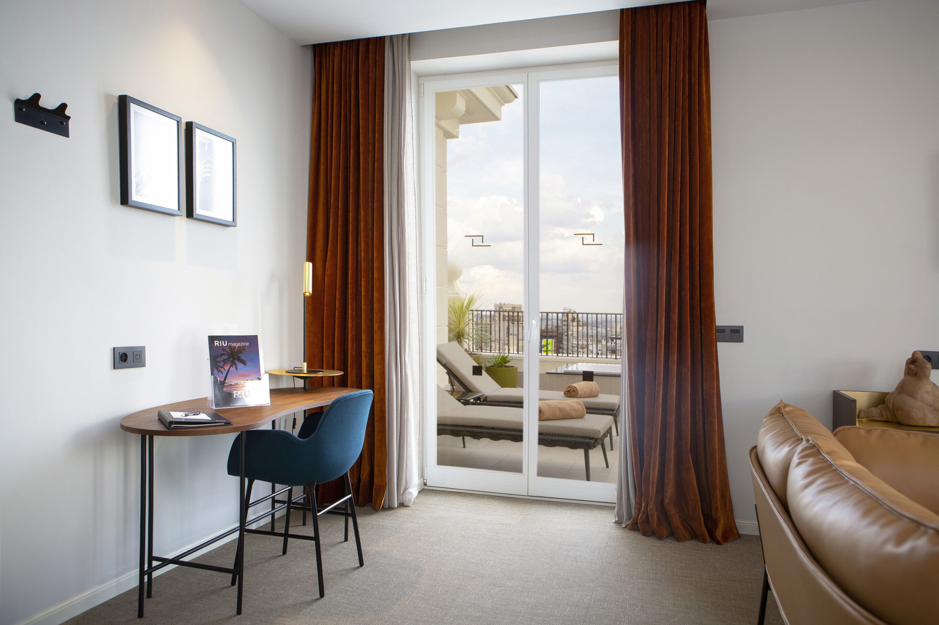 Una habitación de hotel moderna con un escritorio, una silla azul, un sofá de cuero marrón y un balcón con vistas.
